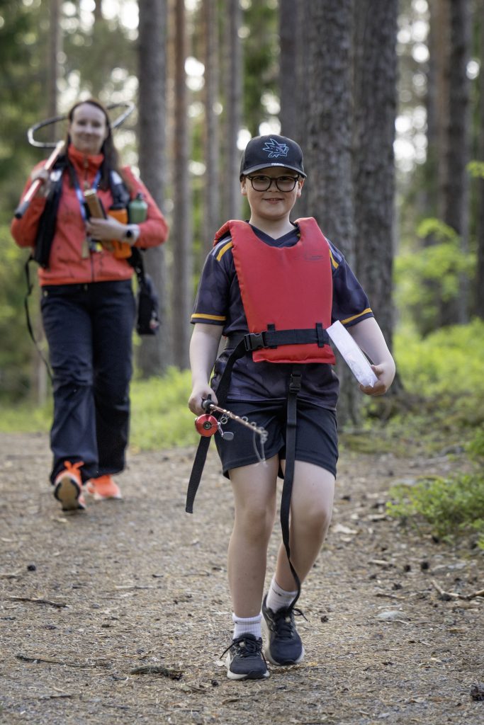 Etualalla Valo kulkee onki kädessään ja punaiset pelastusliivit päällään metsätietä. Taustalla Niina, olallaan haavi ja käsissään 2 vesipulloa.