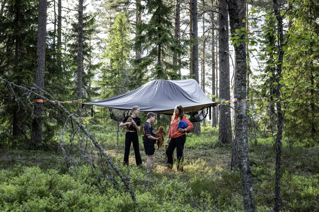 Kaksi lasta ja aikuinen nauravat teltan edessä metsässä. Teltta on ripustettu kolmen puun keskelle, reilu metrin korkeuteen.